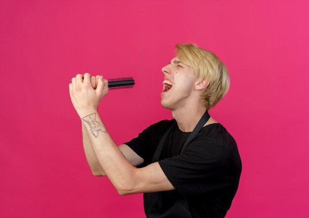 Homme de coiffeur professionnel en tablier tenant une brosse à cheveux en l'utilisant comme microphone en criant ou en chantant