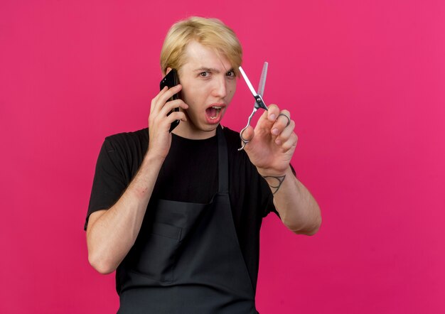 Homme de coiffeur professionnel en tablier tenant des ciseaux parler au téléphone mobile souriant joyeusement