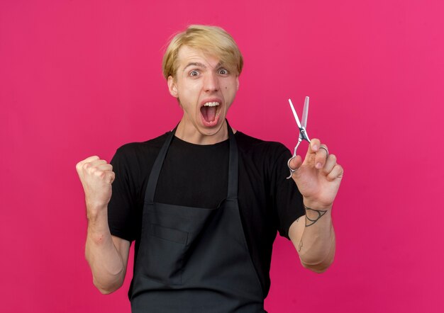 Homme de coiffeur professionnel en tablier tenant des ciseaux serrant le poing se réjouissant fou heureux