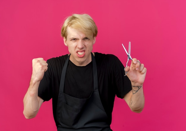 Homme de coiffeur professionnel en tablier tenant des ciseaux serrant le poing avec le visage en colère