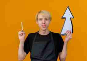 Photo gratuite homme de coiffeur professionnel en tablier tenant une flèche blanche et des ciseaux à l'avant souriant debout sur un mur orange