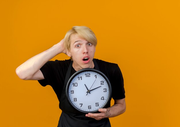 Homme de coiffeur professionnel en tablier tenant horloge murale à l'avant d'être confondu avec la main sur sa tête debout sur un mur orange