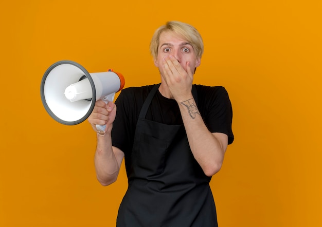 Homme de coiffeur professionnel en tablier tenant un mégaphone couvrant la bouche avec la main étant choqué debout sur un mur orange