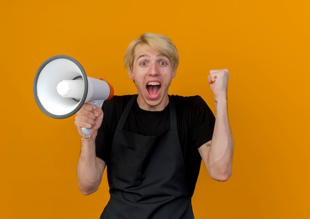 Homme de coiffeur professionnel en tablier tenant le mégaphone serrant le poing se réjouissant heureux et excité debout sur le mur orange