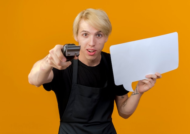 Homme de coiffeur professionnel en tablier tenant un signe de bulle de discours vierge pointant avec tondeuse à l'avant souriant debout sur un mur orange