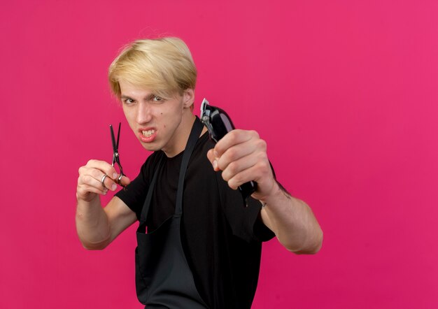 Homme de coiffeur professionnel en tablier tenant tondeuse et ciseaux avec visage en colère