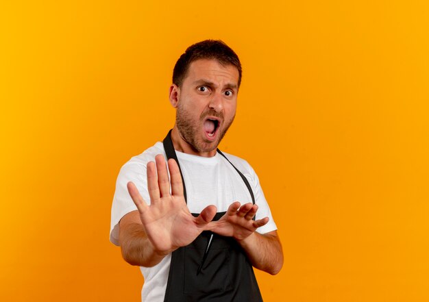 Homme de coiffeur en tablier à l'avant en faisant un geste de défense avec les mains debout sur un mur orange