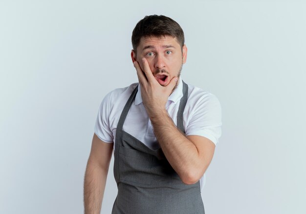 Homme de coiffeur en tablier couvrant la bouche avec la main étant choqué debout sur un mur blanc