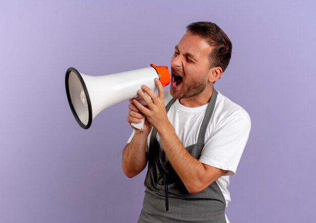 Photo gratuite homme de coiffeur en tablier criant au mégaphone avec une expression agressive debout sur un mur violet