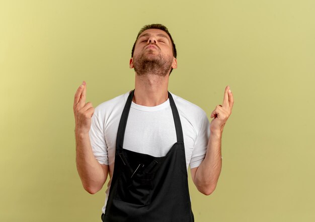 Homme de coiffeur en tablier faisant souhait souhaitable croisant les doigts avec les yeux fermés debout sur un mur lumineux