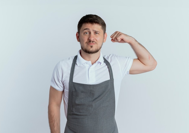 Homme de coiffeur en tablier levant le poing avec une expression confiante, concept gagnant debout sur un mur blanc