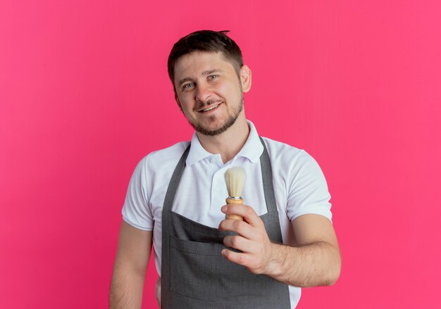 Homme de coiffeur en tablier montrant le pinceau de rasage lookign at camera avec sourire sur le visage debout sur fond rose