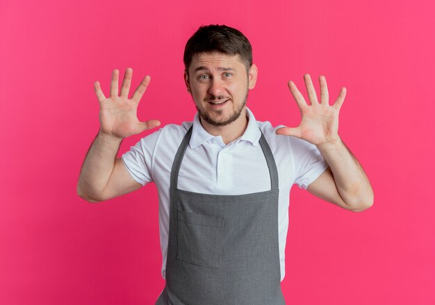 Homme de coiffeur en tablier montrant et pointant vers le haut avec les doigts numéro dix souriant debout sur fond rose