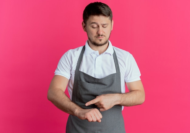 Homme de coiffeur en tablier pointant sur son bras rappelant l'heure avec un visage sérieux debout sur fond rose