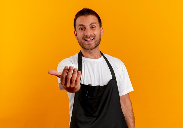 Homme de coiffeur en tablier à la recherche de l'avant invitant à entrer avec main souriant sympathique debout sur un mur orange