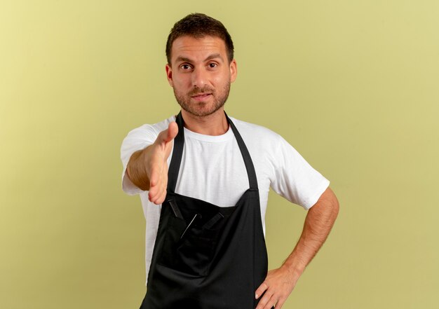 Homme de coiffeur en tablier à la recherche de l'avant souriant offre amicale main salutation debout sur mur léger