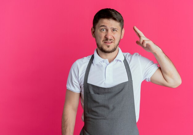 Homme de coiffeur en tablier regardant la caméra avec un sourire sceptique en levant le bras debout sur fond rose