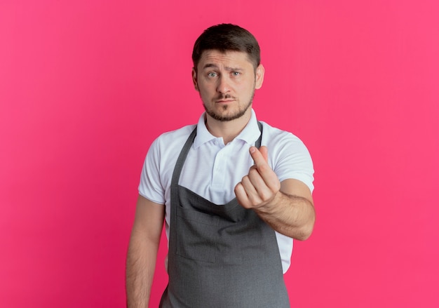 Homme de coiffeur en tablier se frottant les doigts faisant de l'argent geste demandant de l'argent debout sur fond rose