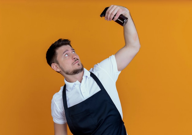 Homme de coiffeur en tablier se rafraîchissant avec un spray debout sur fond orange