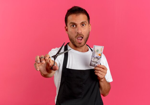 Homme de coiffeur en tablier tenant des ciseaux et de l'argent à l'avant inquiet debout sur le mur rose