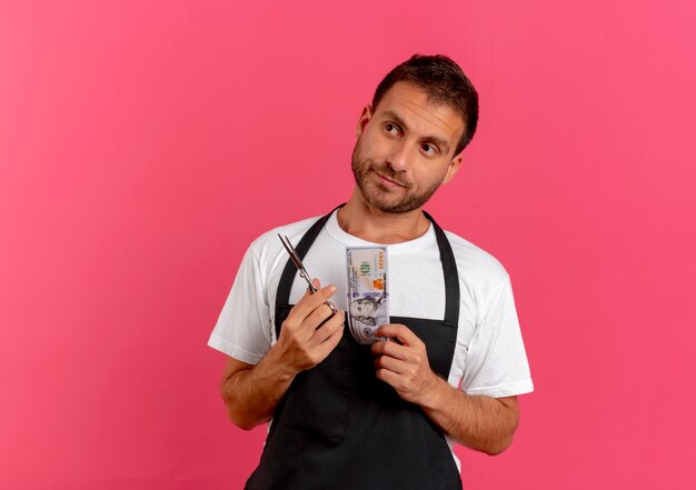 Homme de coiffeur en tablier tenant des ciseaux et de l'argent à côté avec une expression sceptique debout sur un mur rose