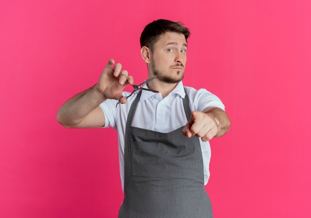 Homme de coiffeur en tablier tenant des ciseaux pointant avec l'index avec une expression confiante debout sur un mur rose