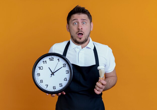 Homme de coiffeur en tablier tenant horloge murale et blaireau étonné et surpris debout sur le mur orange