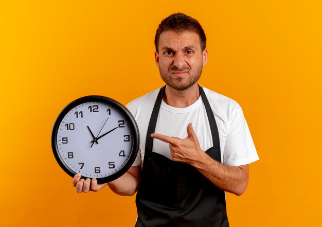 Homme de coiffeur en tablier tenant horloge murale pointant avec le doigt vers elle à l'avant mécontent de se tenir debout sur un mur orange