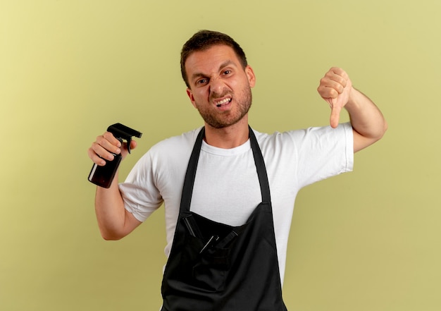 Homme de coiffeur en tablier tenant la machine de coupe de cheveux à mécontent montrant les pouces vers le bas debout sur un mur léger