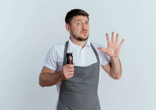 Homme de coiffeur en tablier tenant la machine de coupe de cheveux montrant le numéro cinq à la caméra atr confus debout sur fond blanc