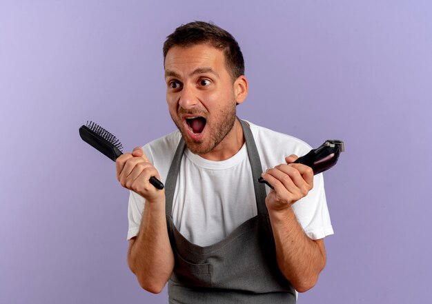 Homme de coiffeur en tablier tenant une machine à couper les cheveux et une brosse à cheveux en criant à la surprise debout sur un mur violet