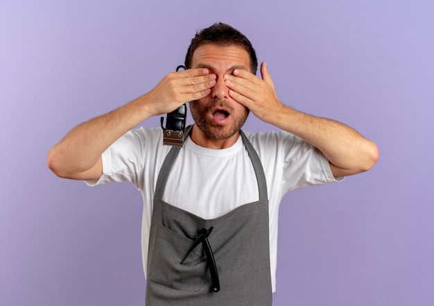 Homme de coiffeur en tablier tenant une machine à couper les cheveux peur couvrant les yeux avec les mains debout sur le mur violet