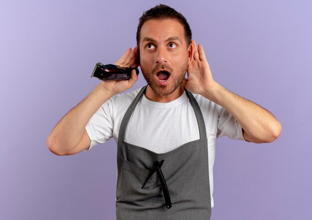 Homme de coiffeur en tablier tenant une machine à couper les cheveux se tenant la main près des oreilles en essayant d'écouter à la surprise debout sur le mur violet