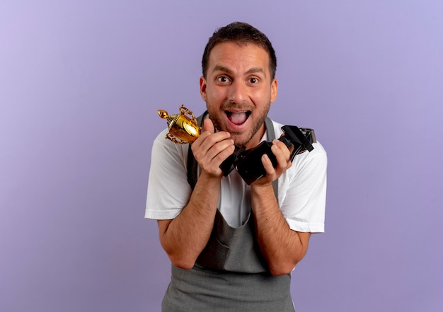 Homme de coiffeur en tablier tenant une machine à couper les cheveux et un trophée à l'avant heureux et excité debout sur un mur violet