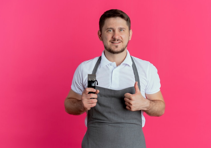 Photo gratuit homme de coiffeur en tablier tenant tondeuse à barbe montrant les pouces vers le haut en souriant regardant la caméra debout sur fond rose