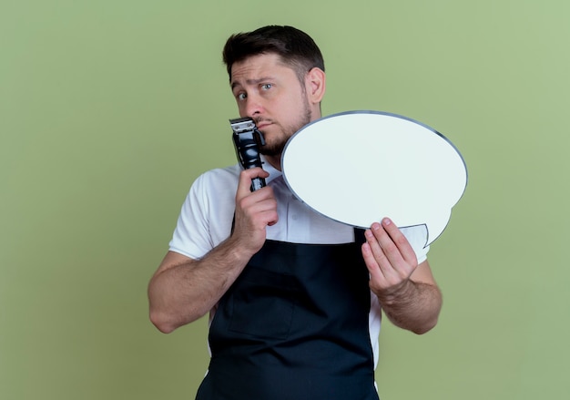 Homme de coiffeur en tablier tenant tondeuse à barbe et signe de bulle de discours vide perplexe debout sur le mur vert