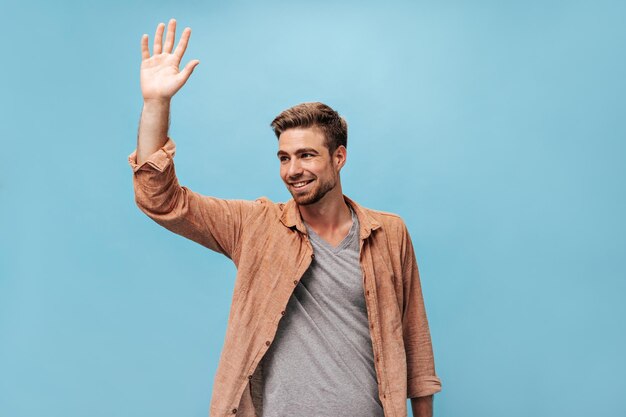 Homme avec une coiffure et une barbe élégantes en T-shirt et chemise marron à manches longues regardant loin souriant et saluant sur fond bleu