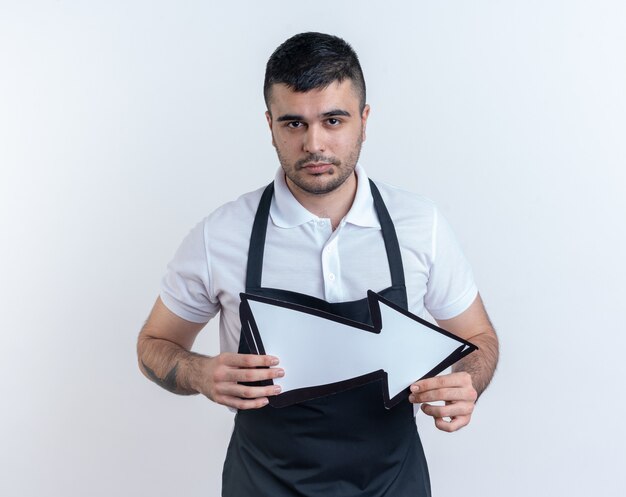 Homme de coiffure en tablier tenant une flèche regardant la caméra avec une expression sérieuse et confiante, debout sur fond blanc