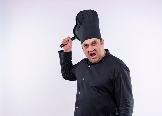 Homme en colère chef cuisinier portant l'uniforme noir et chapeau de cuisinier balançant une casserole avec une expression agressive debout sur fond blanc
