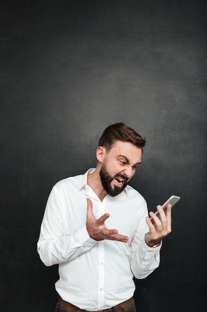 Homme en colère criant d'être irrité tout en regardant sur smartphone dans sa main sur gris foncé