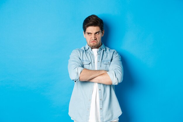 Un homme en colère croise les bras sur la poitrine et regarde au loin avec une expression insultée, debout offensé sur fond bleu