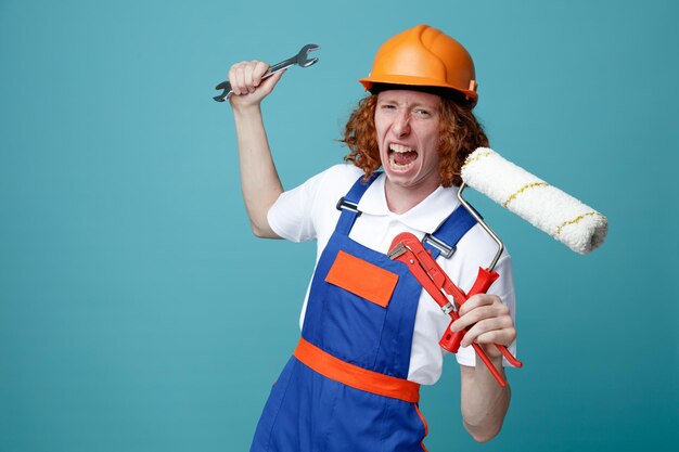 Homme en colère jeune constructeur en uniforme tenant des outils de construction isolés sur fond bleu