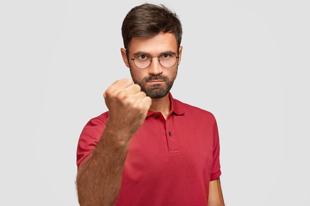 Un homme en colère sérieux montre son poing, prêt pour le combat ou le défi, a une expression sévère, porte un t-shirt rouge décontracté, pose contre un mur blanc. Agressif jeune homme fait des gestes à l'intérieur. Concept de langage corporel