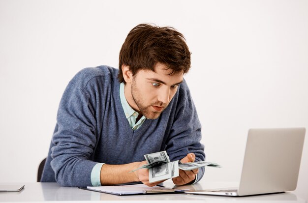 Homme comptant de l'argent avec une expression folle, a gagné beaucoup d'argent, ne peut pas croire qu'il a gagné à la loterie, assis au bureau près d'un ordinateur portable