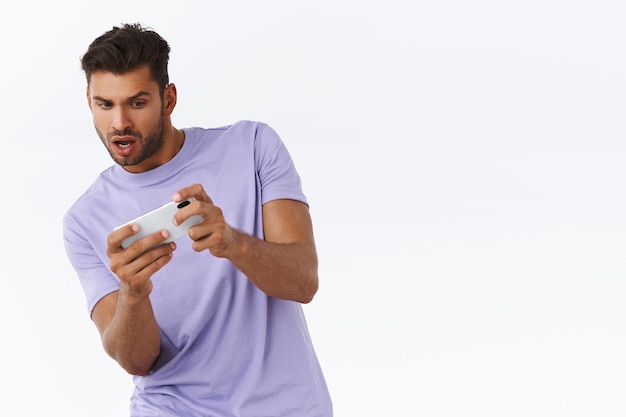 Homme concentré et excité avec un nouveau jeu de course génial, jouant sur un smartphone, inclinant le corps pour déplacer la voiture sur un téléphone portable, regardant le téléphone ravi et diverti