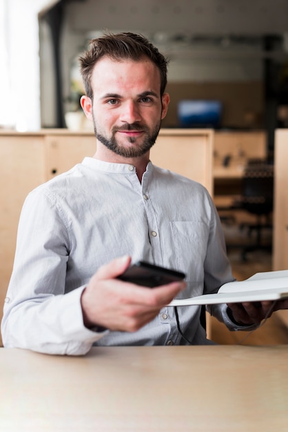 Photo gratuite homme confiant tenant un téléphone cellulaire et son journal en regardant la caméra