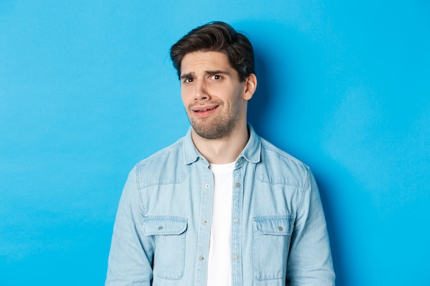 Homme confus et mal à l'aise regardant quelque chose d'étrange ou d'effrayant, grincer des dents d'une mauvaise publicité, debout sur fond bleu