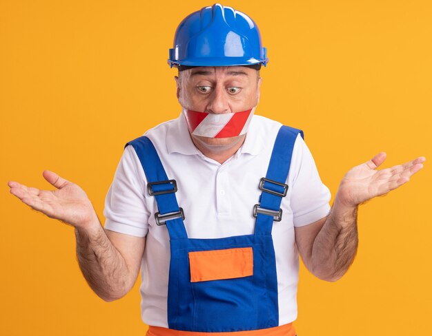 Homme de constructeur adulte anxieux en uniforme couvre la bouche avec du ruban adhésif et tient les mains ouvertes regardant vers le bas isolé sur mur orange