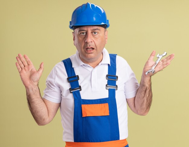 Homme constructeur adulte désemparé en uniforme se tient avec les mains levées tenant la clé isolée sur le mur vert olive