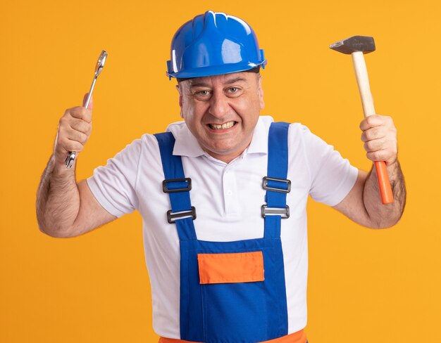 Homme de constructeur adulte excité en uniforme détient une clé et un marteau isolé sur mur orange